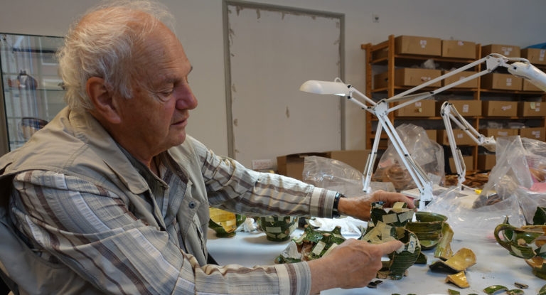 Vrijwiliger Frans aan een tafel vol scherven en speciale lampen. In zijn handen heeft hij scherven vast.