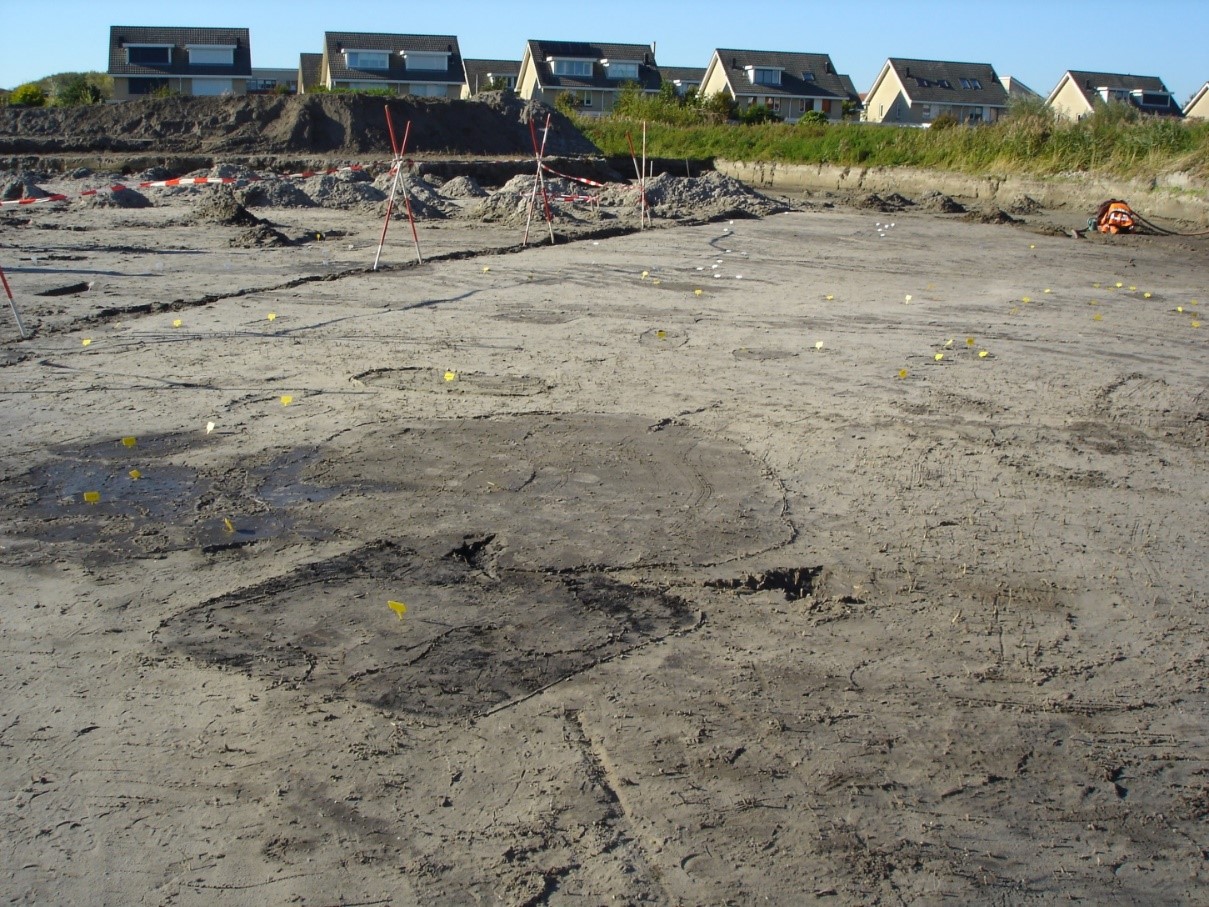 Foto van afvalkuilen bij de opgraving Katwijk de Zanderij. Op de voorgrond zie je een zandvlakte, op de achtergrond staan zes huizen. Er staan geen mensen op de foto