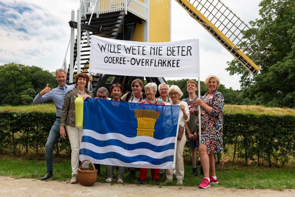 Het team van Streekmuseum Goeree-Overflakkee poseert met hun zelfgemaakte spandoek voor de molen van het museum. Op het spandoek staat de leus 'Wille wete nie beter Goeree-Overflakkee