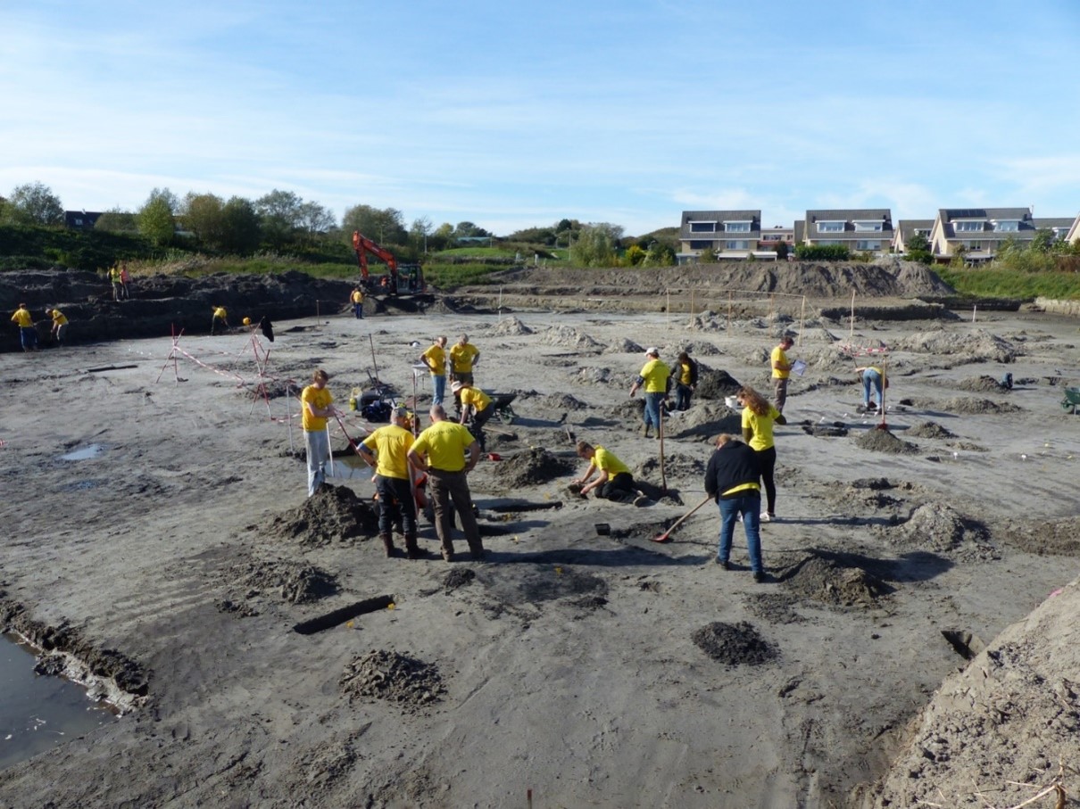 Sfeerbeeld van de opgraving Katwijk de Zanderij. De mensen op de foto hebben gele t-shirts aan en staan of zitten in een zandvlakte. De foto is van een afstand genomen.