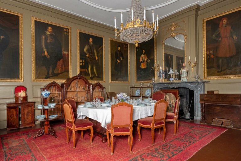 Kamer in Slot Zuylen met geschilderde portretten aan de wand, een kroonluchter aan het plafond en daaronder een gedekte tafel met daaromheen antieke stoelen. Op de grond ligt een rood tapijt.